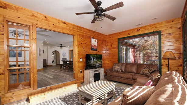 L’intérieur d’un chalet confortable, avec un salon, des murs en bois, un ventilateur de plafond, un téléviseur et une grande fenêtre.
