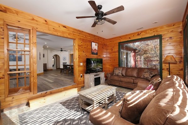 L’intérieur d’un chalet confortable, avec un salon, des murs en bois, un ventilateur de plafond, un téléviseur et une grande fenêtre.
