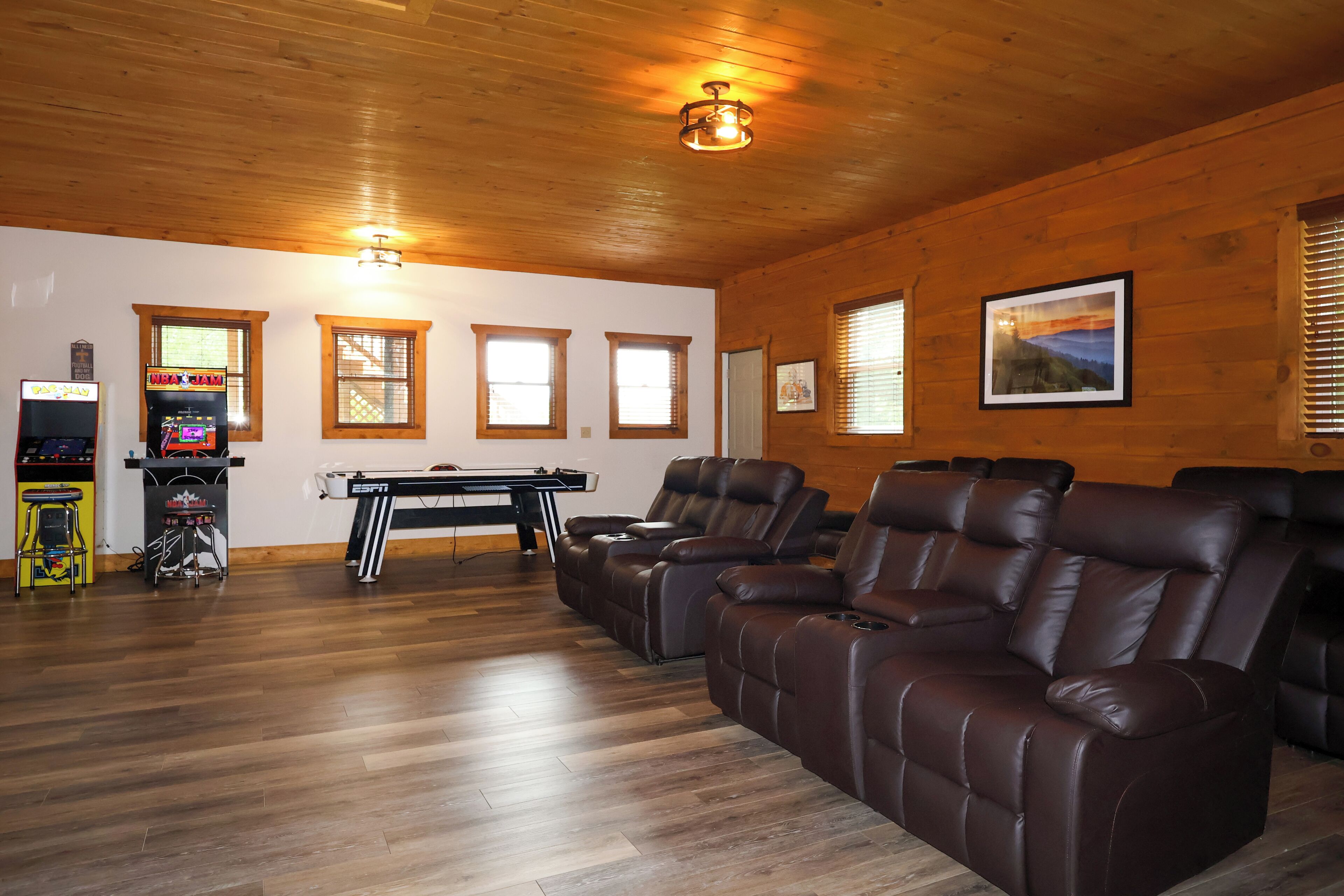 A room with wooden flooring, a piano, leather recliners, and arcade games.