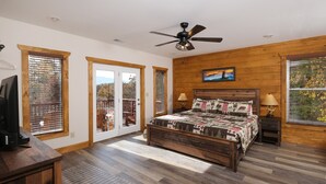 Une chambre avec un lit en bois, un ventilateur de plafond et une vue sur l’extérieur grâce à de grandes fenêtres.