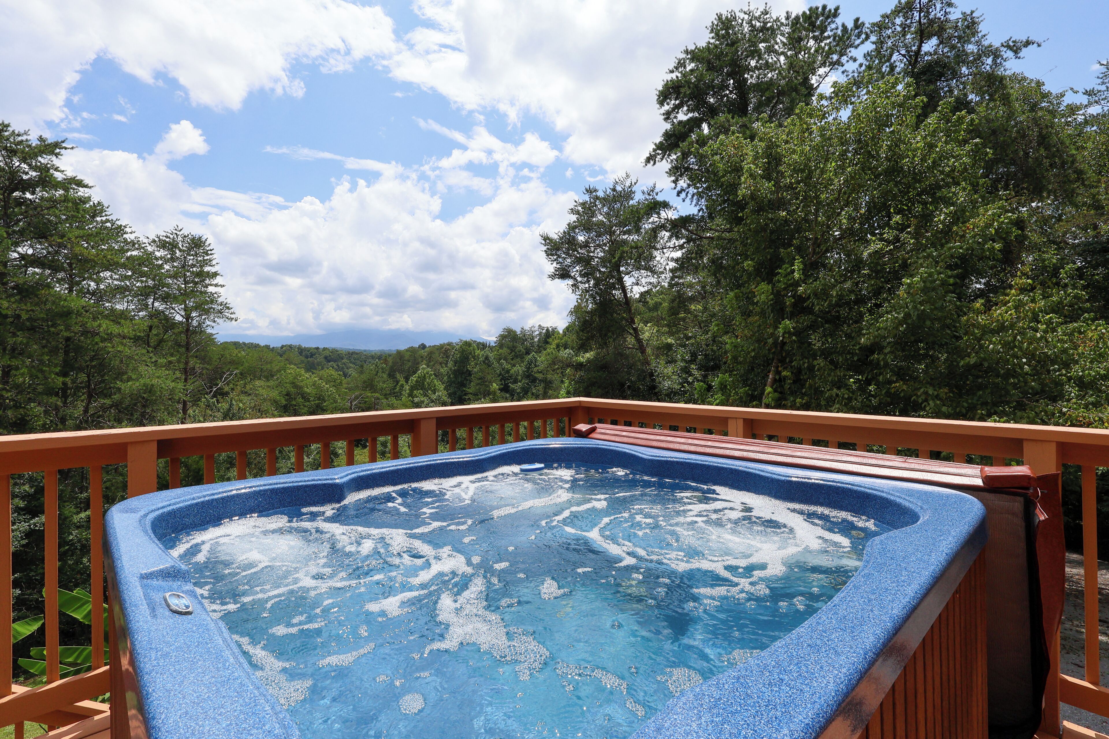 A hot tub surrounded by a wooden deck with a scenic view of trees and mountains in the background.