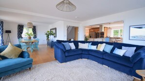 A spacious living room with a blue sectional sofa, a dining area with a table and chairs, and a kitchen in the background. - Ty Ni (New Quay)