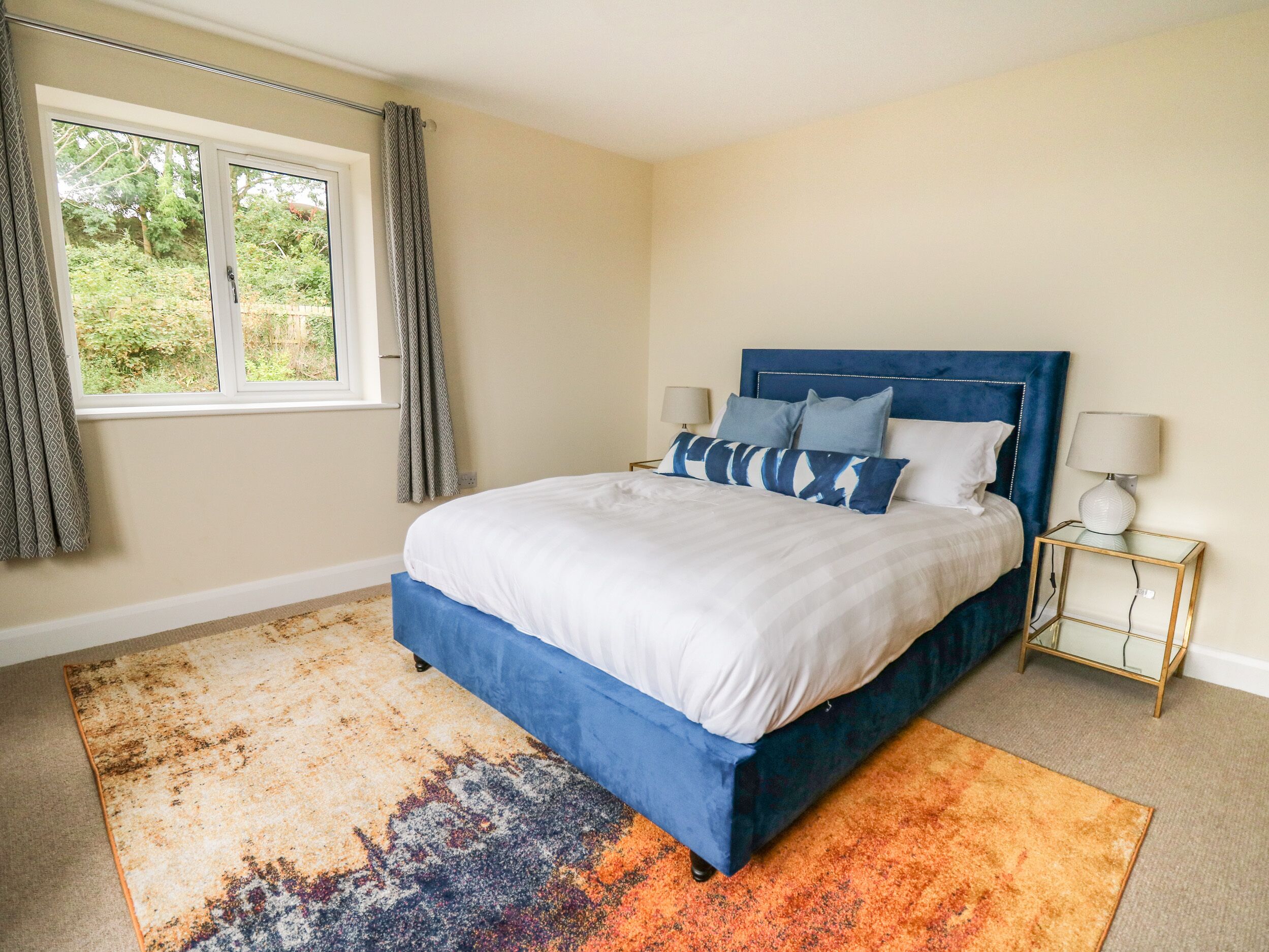 A bedroom with a large bed, a window with curtains, a nightstand with a lamp, and a colorful rug.