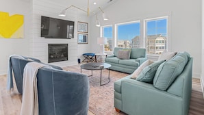 A spacious living room with a fireplace, a large window with a view of buildings, and modern furniture. - Sea Salt & Sand w/ Heated Pool + Near the Beach (Corolla)