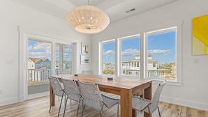 A spacious dining area with a wooden table and wicker chairs, large windows with a view of houses, and a hanging light fixture. - Sea Salt & Sand w/ Heated Pool + Near the Beach (Corolla)