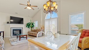 A spacious living room with a fireplace, a dining area with a glass table, and a large window with blinds. - OBX Ridgeview 4 Bedroom Home at Southridge (Nags Head)