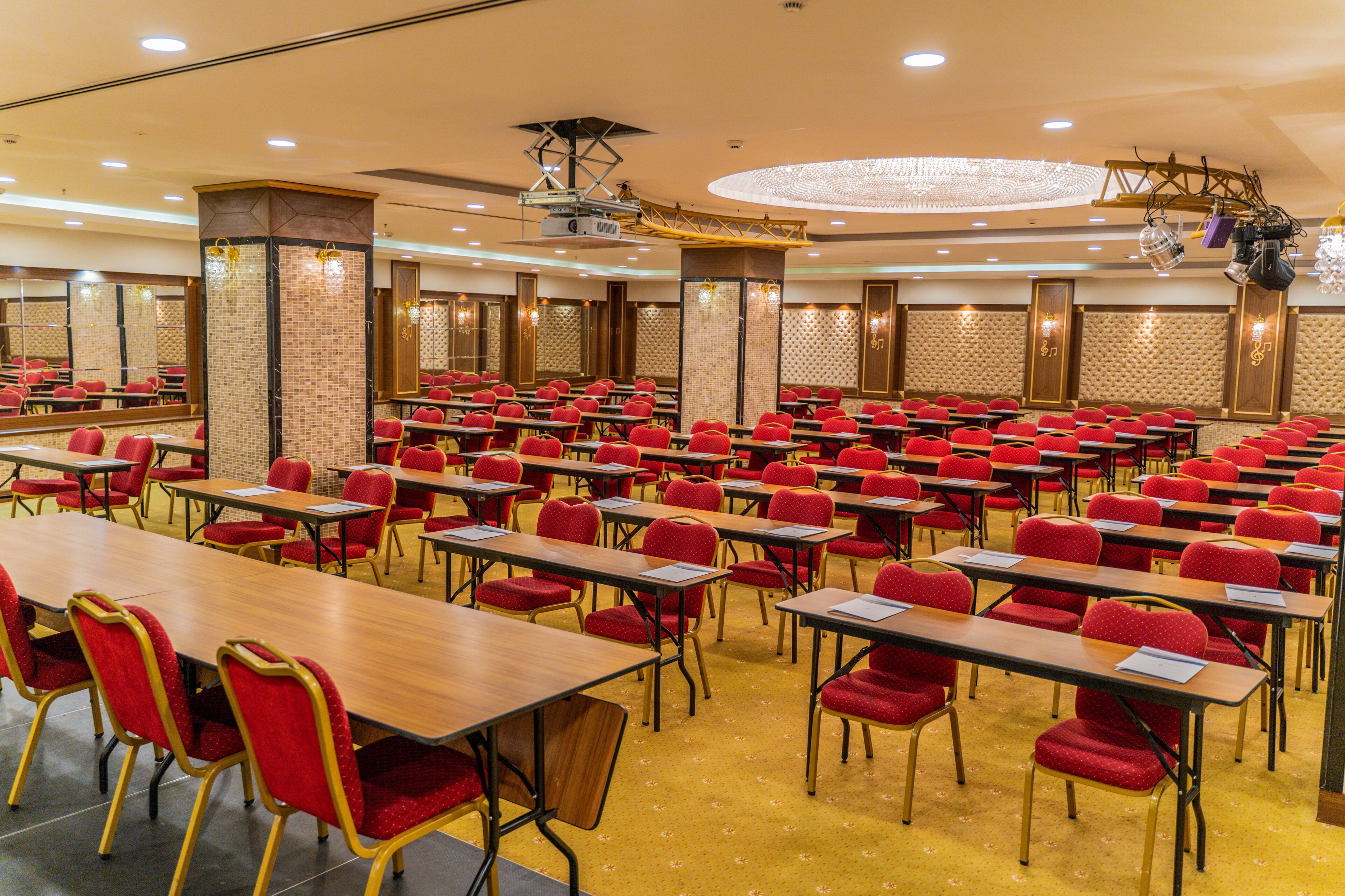 A conference room with rows of tables and chairs, a stage with a projector, and a chandelier.