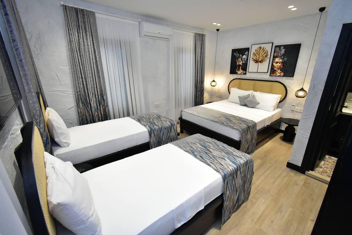 A hotel room with two single beds, a large window with curtains, a wall-mounted artwork, and a small table with a lamp.