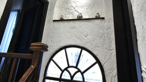 A room with a wooden staircase, a white textured wall, a small shelf with decorative items, and a large arched window. - Otel Gülayvalık (Ayvalik)