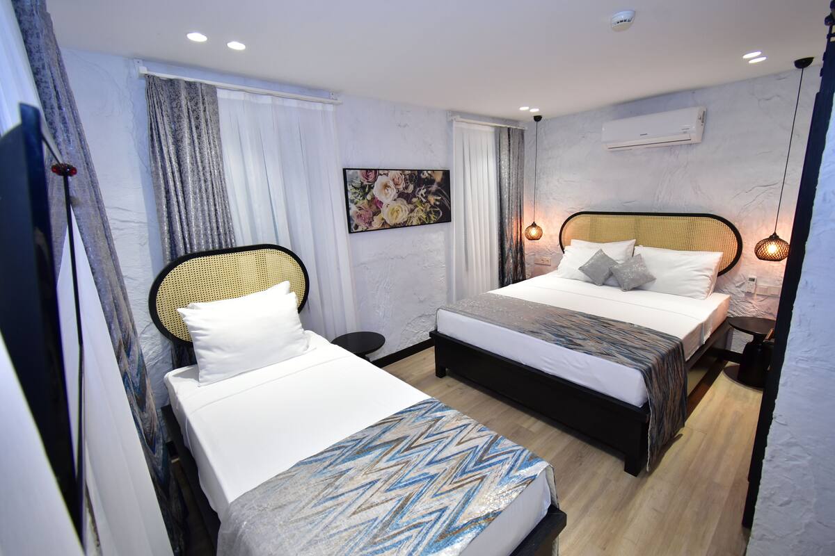 A hotel room with two beds, a large window with curtains, a wall-mounted air conditioner, and a painting.