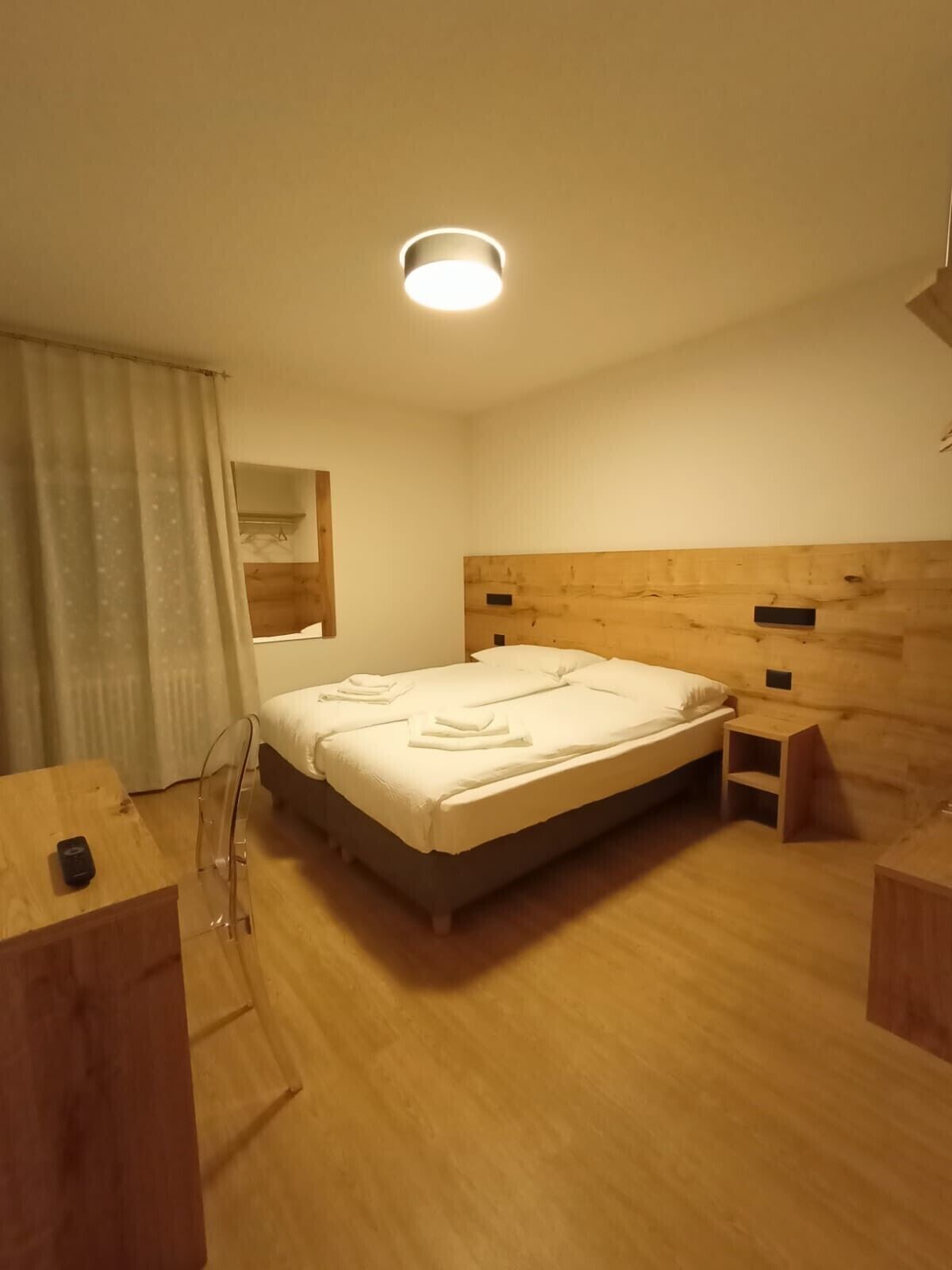 Una habitación de hotel con cabecera de madera, cama con ropa blanca, una mesita de noche de madera, un escritorio de madera y un espejo en la pared.