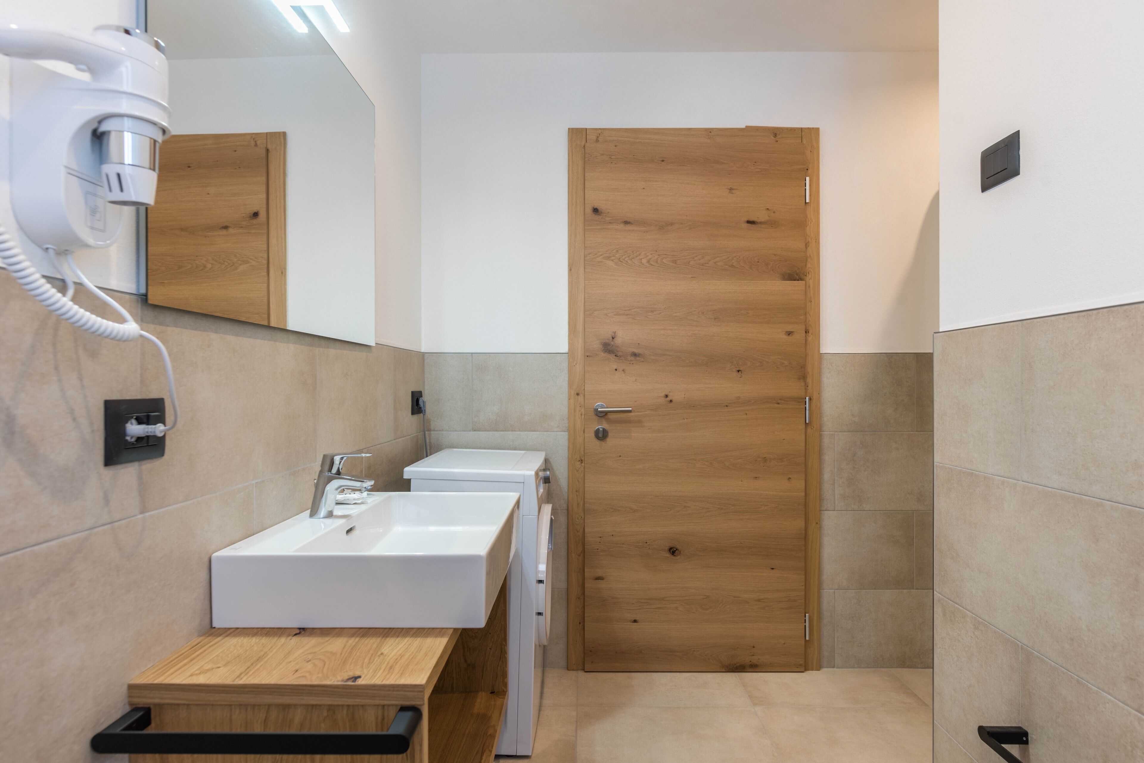 A modern bathroom with a wooden door, a white sink, and a hairdryer on a wall-mounted station.