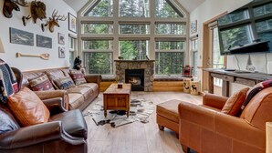 A cozy living room with leather sofas, a stone fireplace, and large windows overlooking a forest. - Call to the Wild in Canmore! (Canmore)