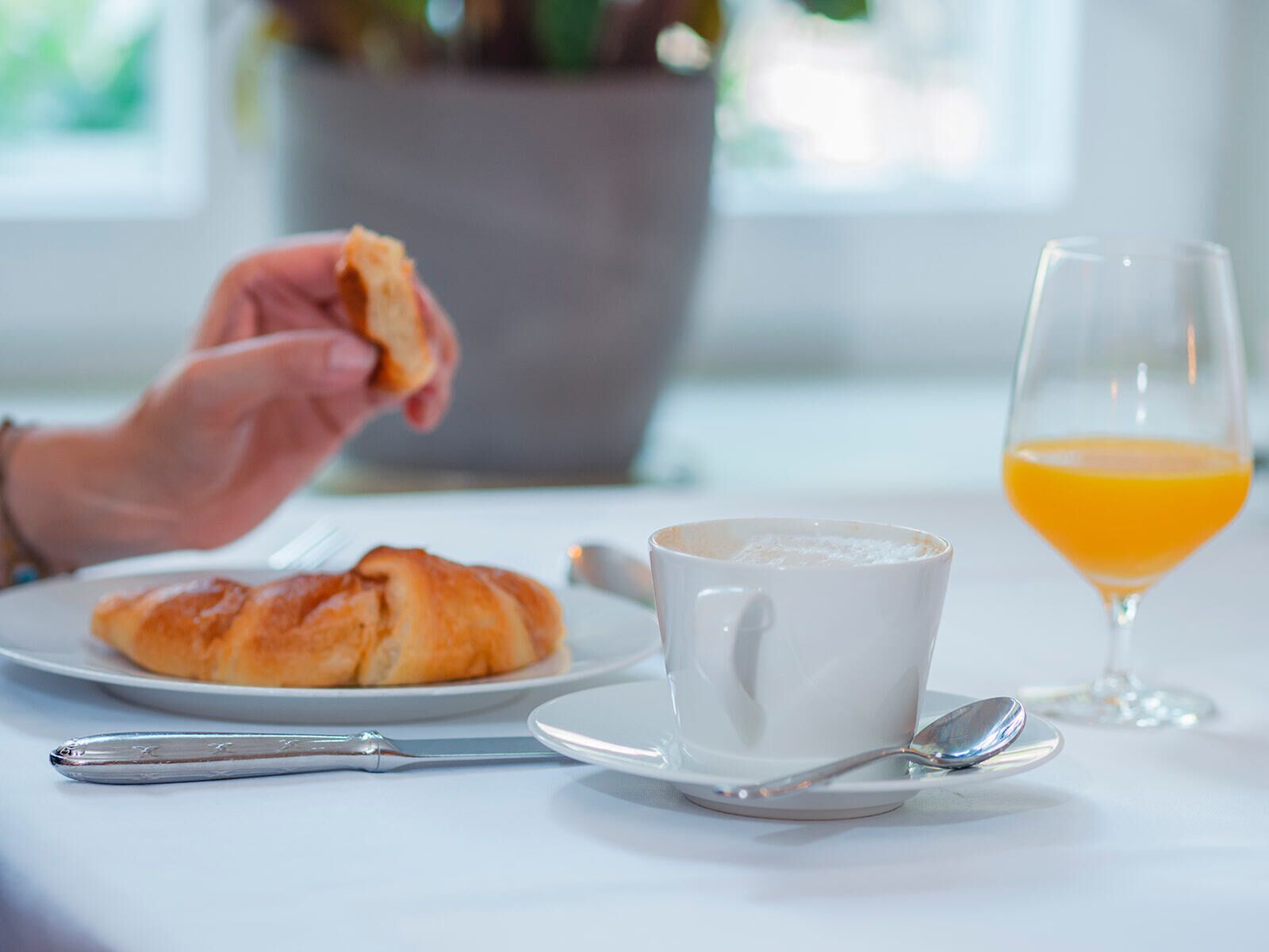 A breakfast setting with a croissant, a cup of coffee, a glass of orange juice, and a spoon on a white table.
