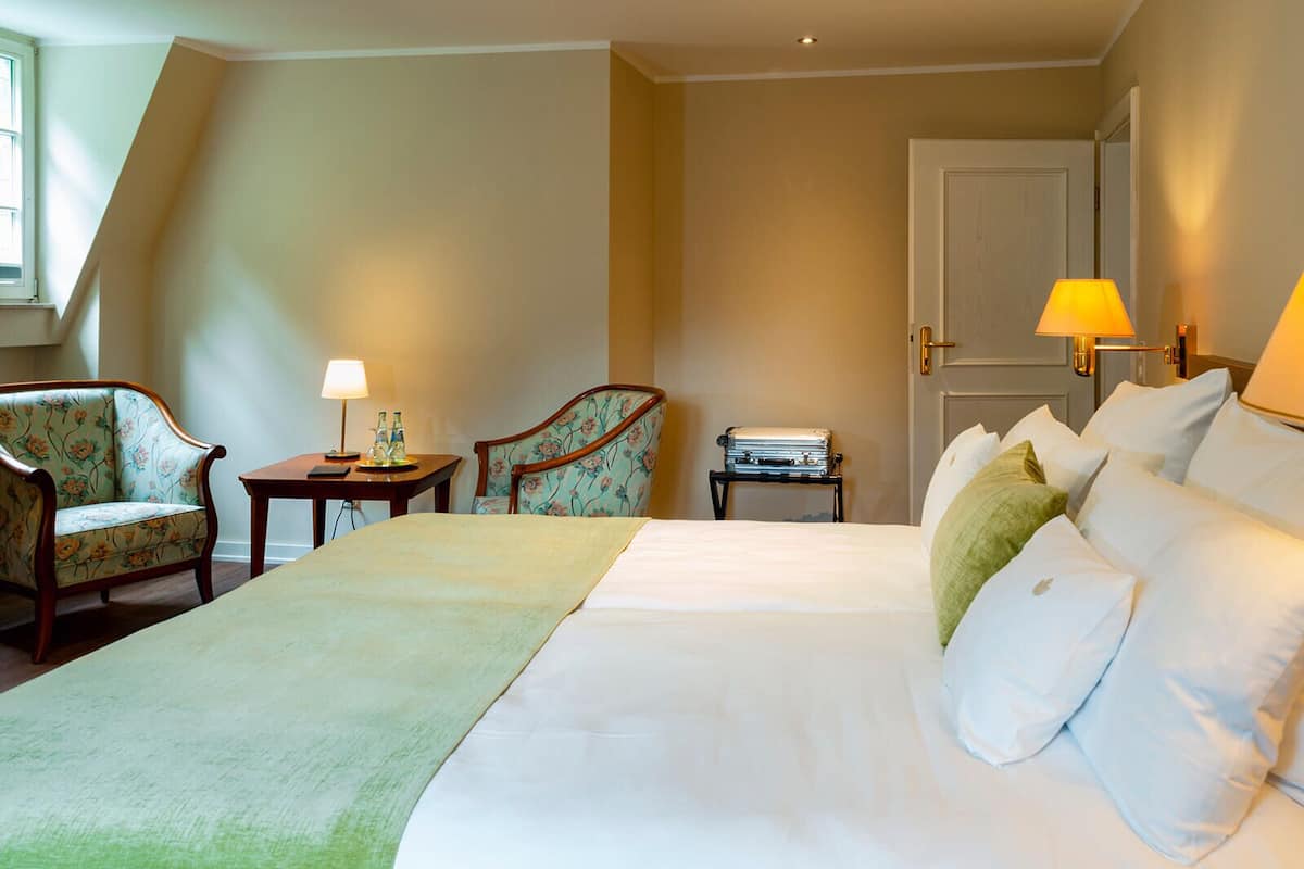Een hotelkamer met een groot bed, twee fauteuils, een bijzettafeltje met een lamp, en een koffer op een standaard.