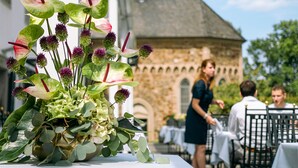 A table with a floral arrangement featuring green leaves and purple flowers, set outdoors with a stone building and a church tower in the background.