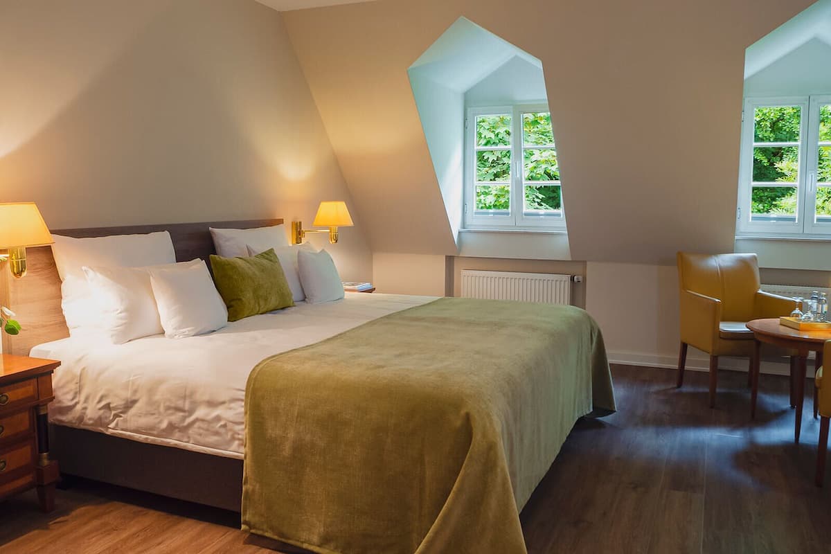 Een slaapkamer met een groot bed, een bureau en een stoel. Er is een raam met uitzicht op groen.