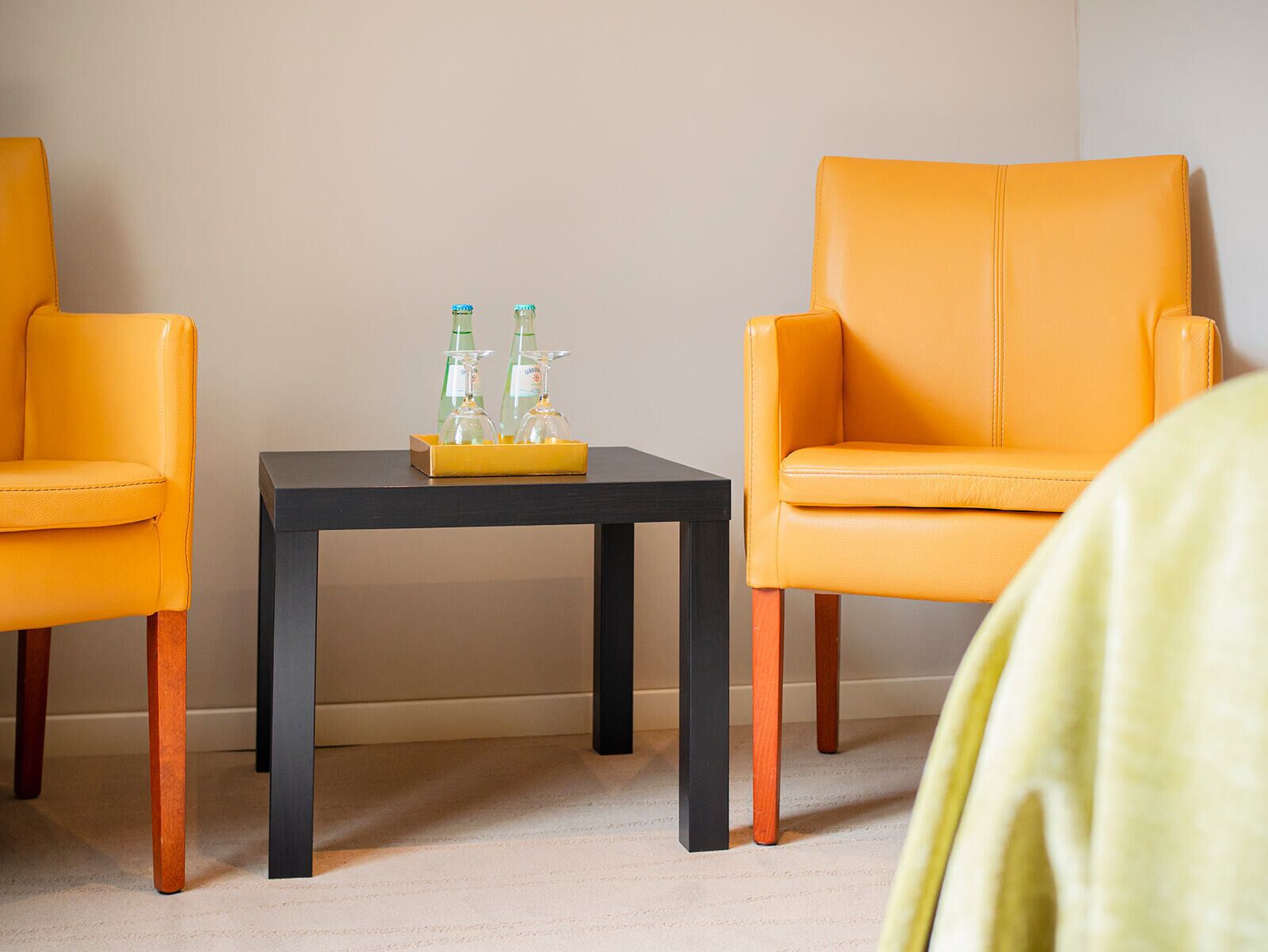 Une pièce avec deux fauteuils en cuir jaune, une table noire sur laquelle se trouve un plateau avec deux bouteilles et des verres, et un mur sobre.