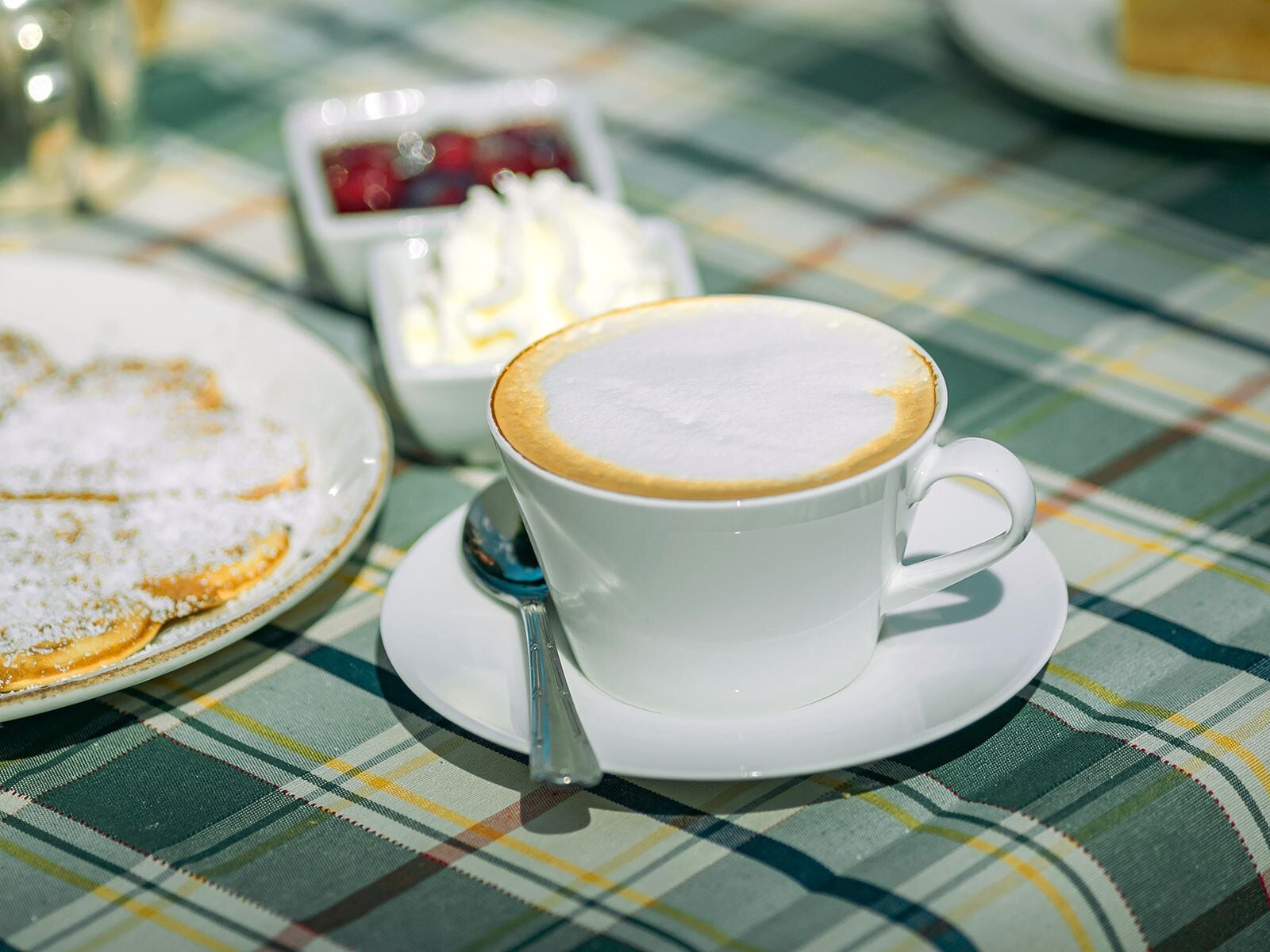 Una taza de café con una capa cremosa sobre un platillo blanco, una cuchara y un plato con un postre sobre un mantel escocés.