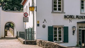 A white building with green shutters, labeled "HOTEL ALIENBERGER HOF," featuring a cobblestone entrance and outdoor seating area.