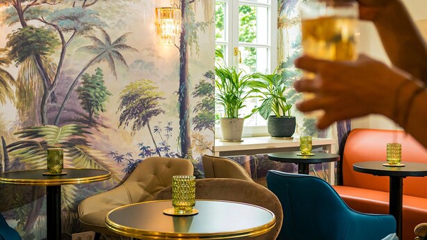 A room with a mural of tropical trees, a window with potted plants, and a table with a glass.