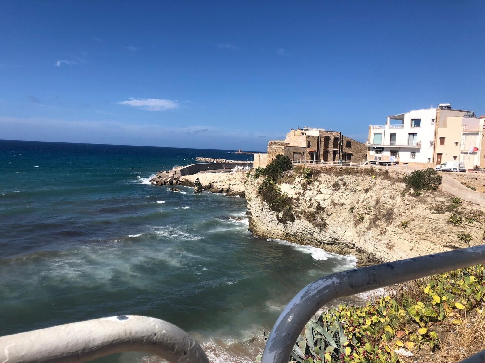 Vista costeira com linha de costa rochosa, um cais que se estende para o mar e edifícios próximos à costa.