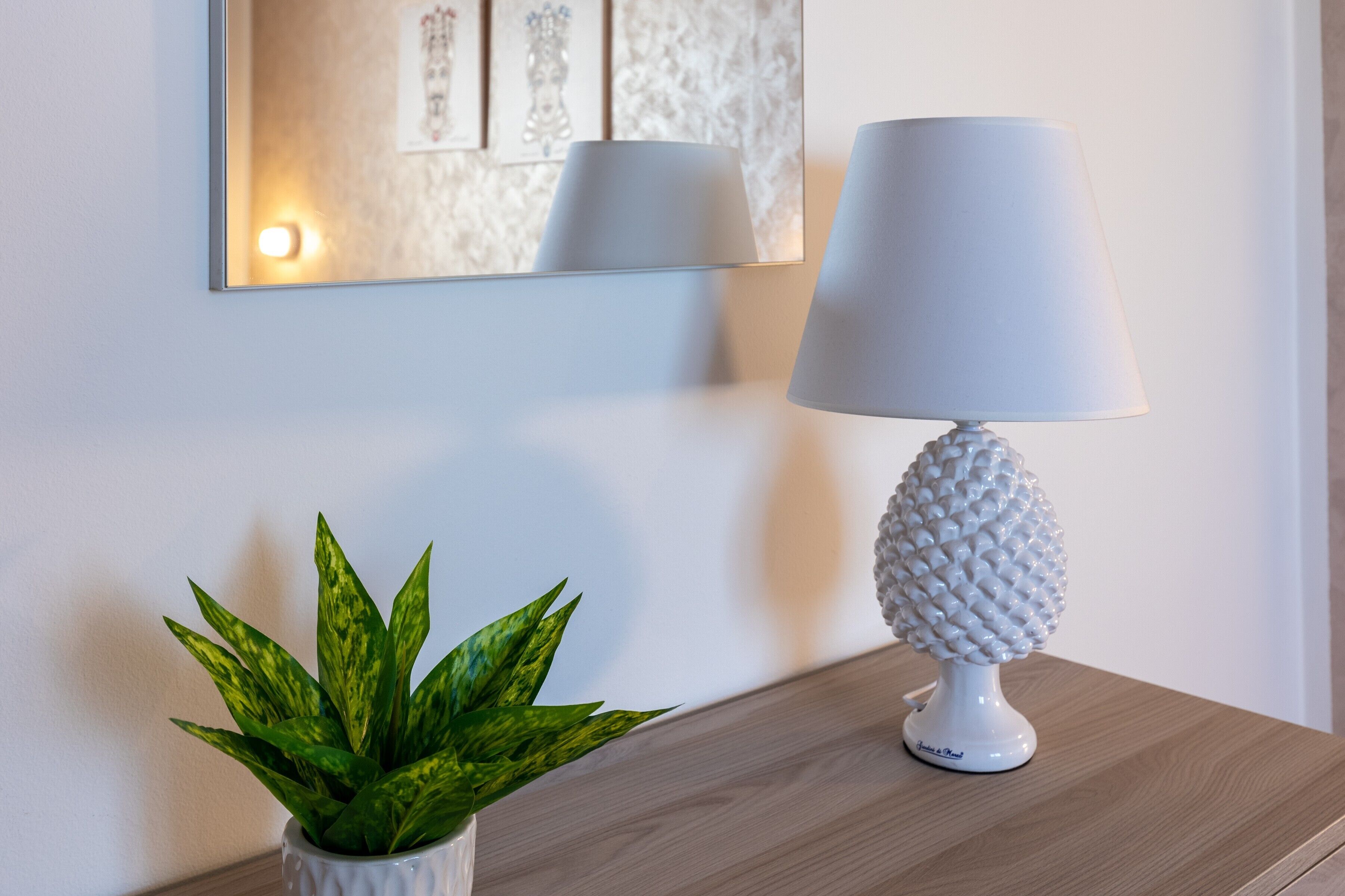 Une lampe blanche à la base texturée, posée sur une surface en bois, une plante en pot aux feuilles panachées et un miroir mural qui reflète la lampe et une œuvre d’art encadrée.