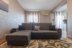 Uma sala de estar com um sofá modular cinza, um tapete estampado e uma janela grande com cortinas.