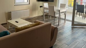 A modern living room with a sofa, a TV mounted on the wall, a dining area with chairs, and a large window. - Cirpan Apart Otel (Enez)