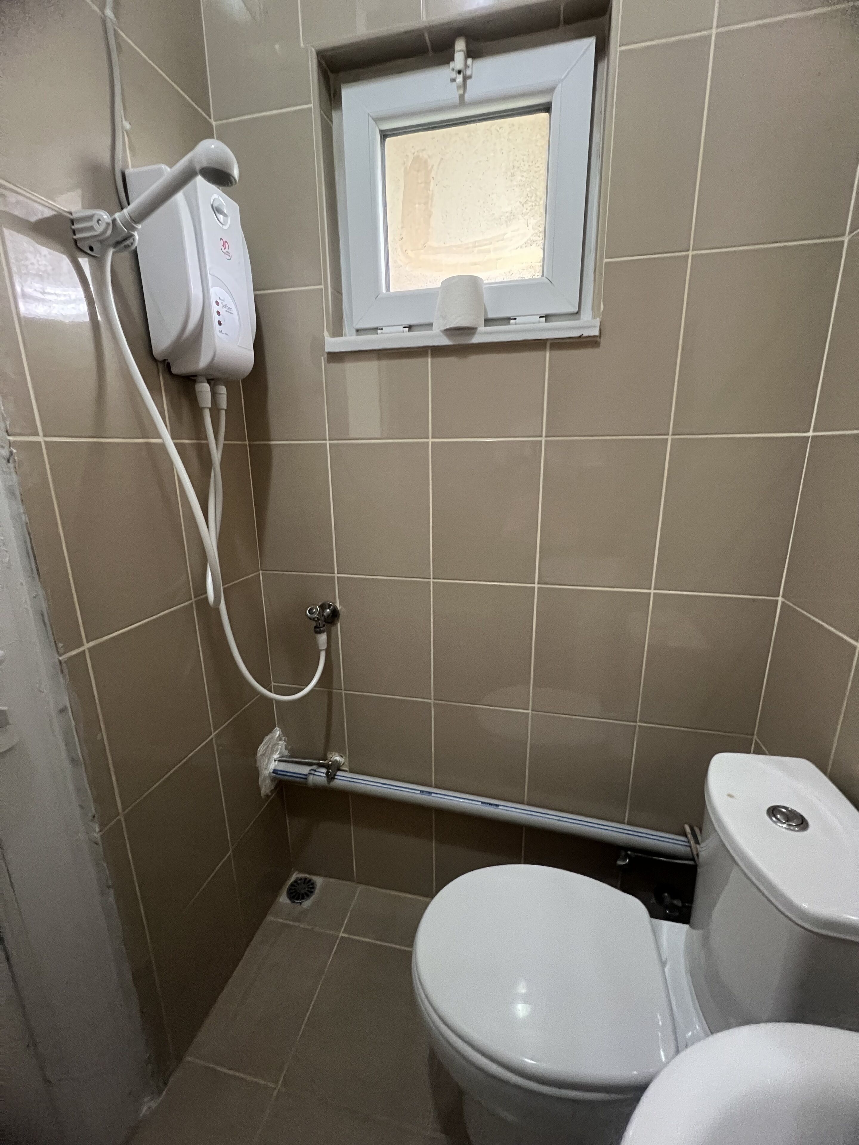 Een kleine betegelde badkamer met toilet, raam en douchekraan.