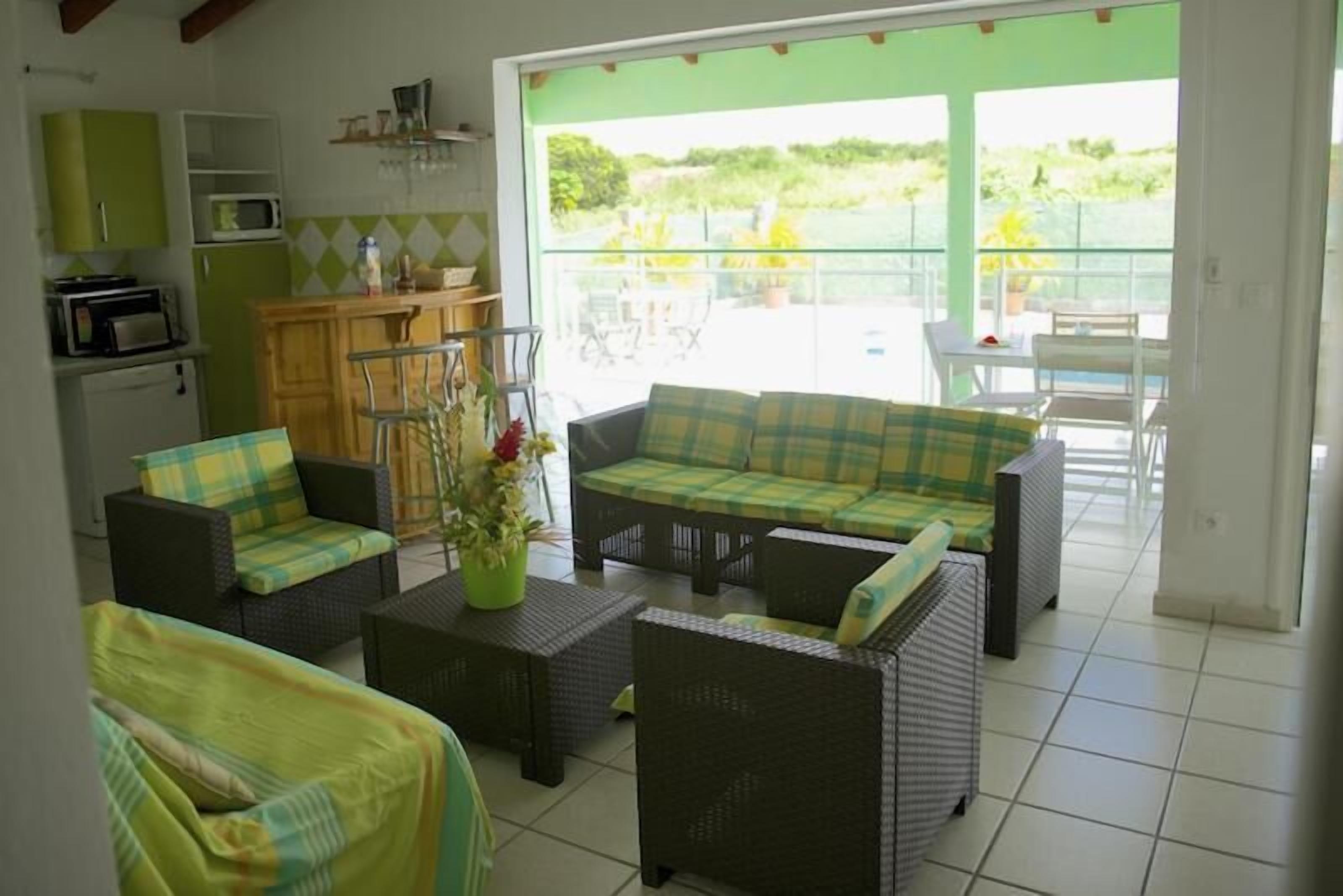 A cozy living area with wicker furniture, a green sofa, and a coffee table. A kitchen with a microwave and cabinets is visible in the background.