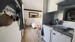 A compact kitchen with a sink, stove, and white cabinets. There is a bed with a patterned bedspread in the adjacent room. - Air-conditioned studio with parking and close to the sea. (Carqueiranne)