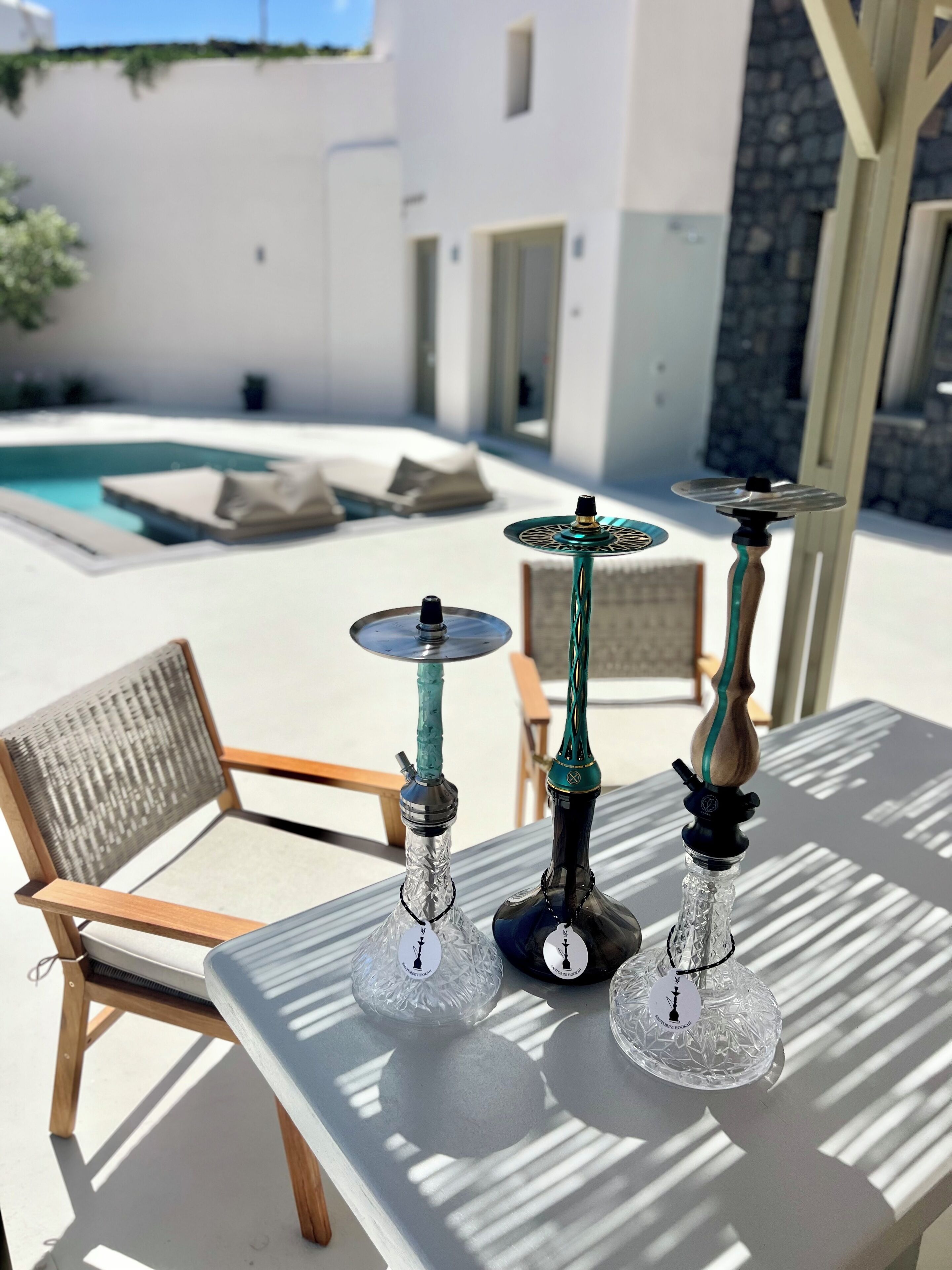 Mesa de vidrio con tres cachimbas y una silla en un espacio exterior soleado con piscina y paredes de piedra al fondo.