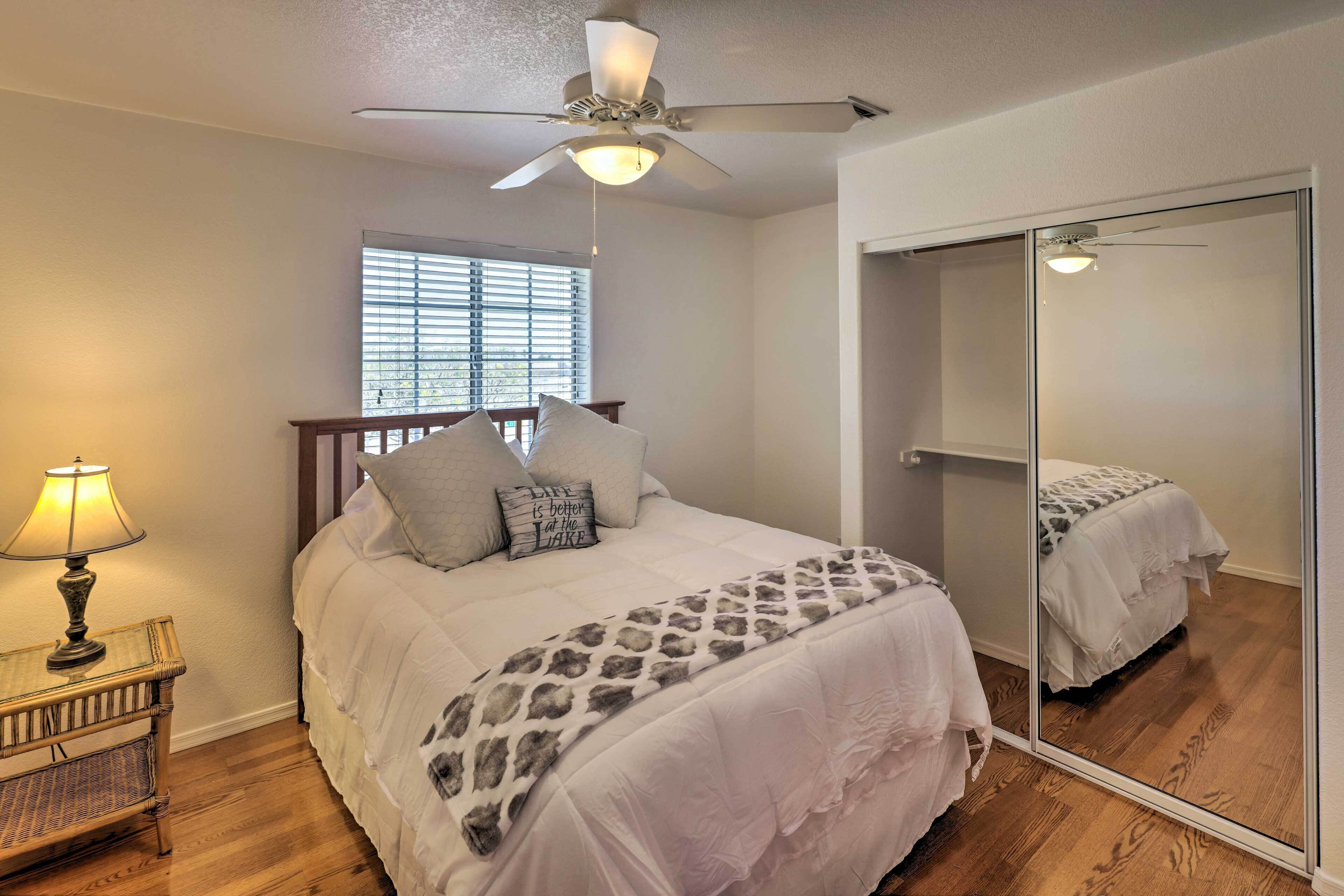 A bedroom with a bed, a ceiling fan, a window with blinds, a bedside table with a lamp, and a large mirror.