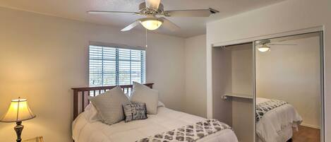 A bedroom with a bed, a ceiling fan, a window with blinds, a bedside table with a lamp, and a large mirror.