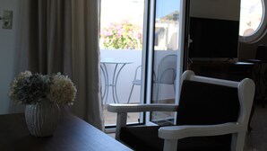 A room with a white chair, a small table, and a vase with flowers. There is a window with curtains, and a view of the outside. - Dimokritos Apartment Pitsidia (Pitsidia)