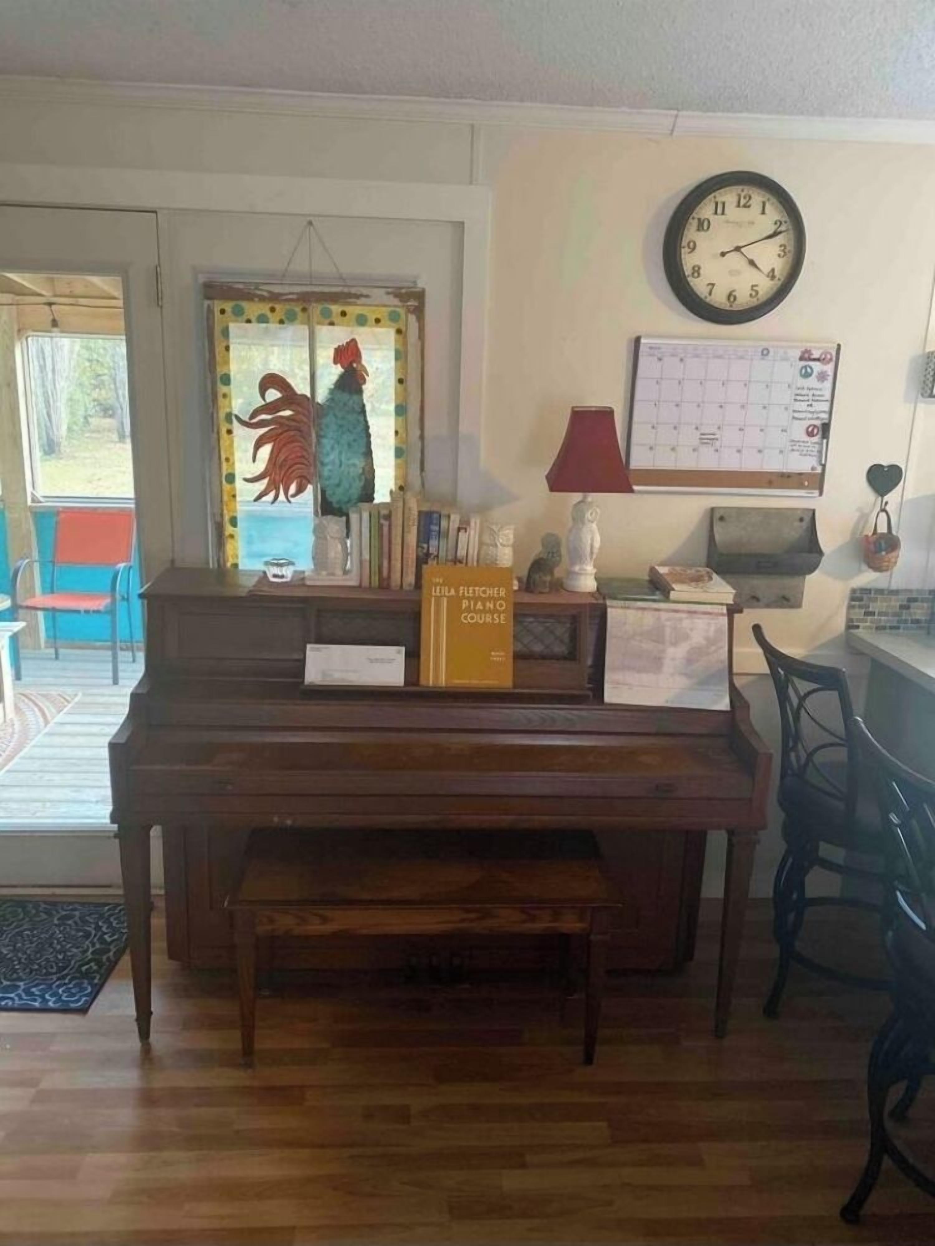 A living room with a wooden piano, a wall clock, a calendar, and a rooster decoration.