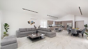 Un salon moderne avec un canapé gris, un pouf et une table basse, un coin repas avec des chaises grises, et une cuisine en arrière-plan.