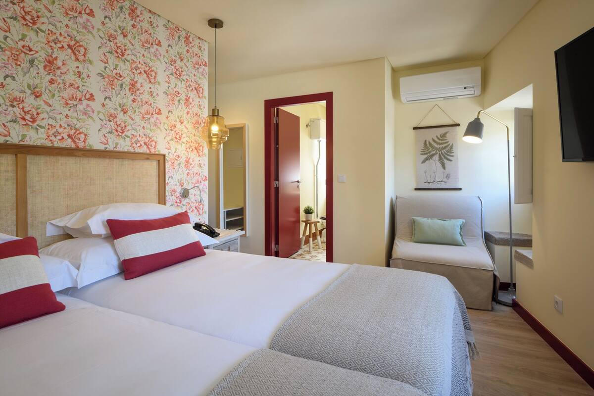 A hotel room with a large bed, a desk, a chair, a small table, and a wall-mounted TV.