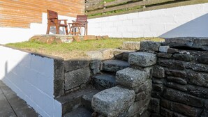 A rustic outdoor seating area with wooden chairs and a table, a stone wall, and a white building with a wooden roof. - Dolphin Cottage (Banff)