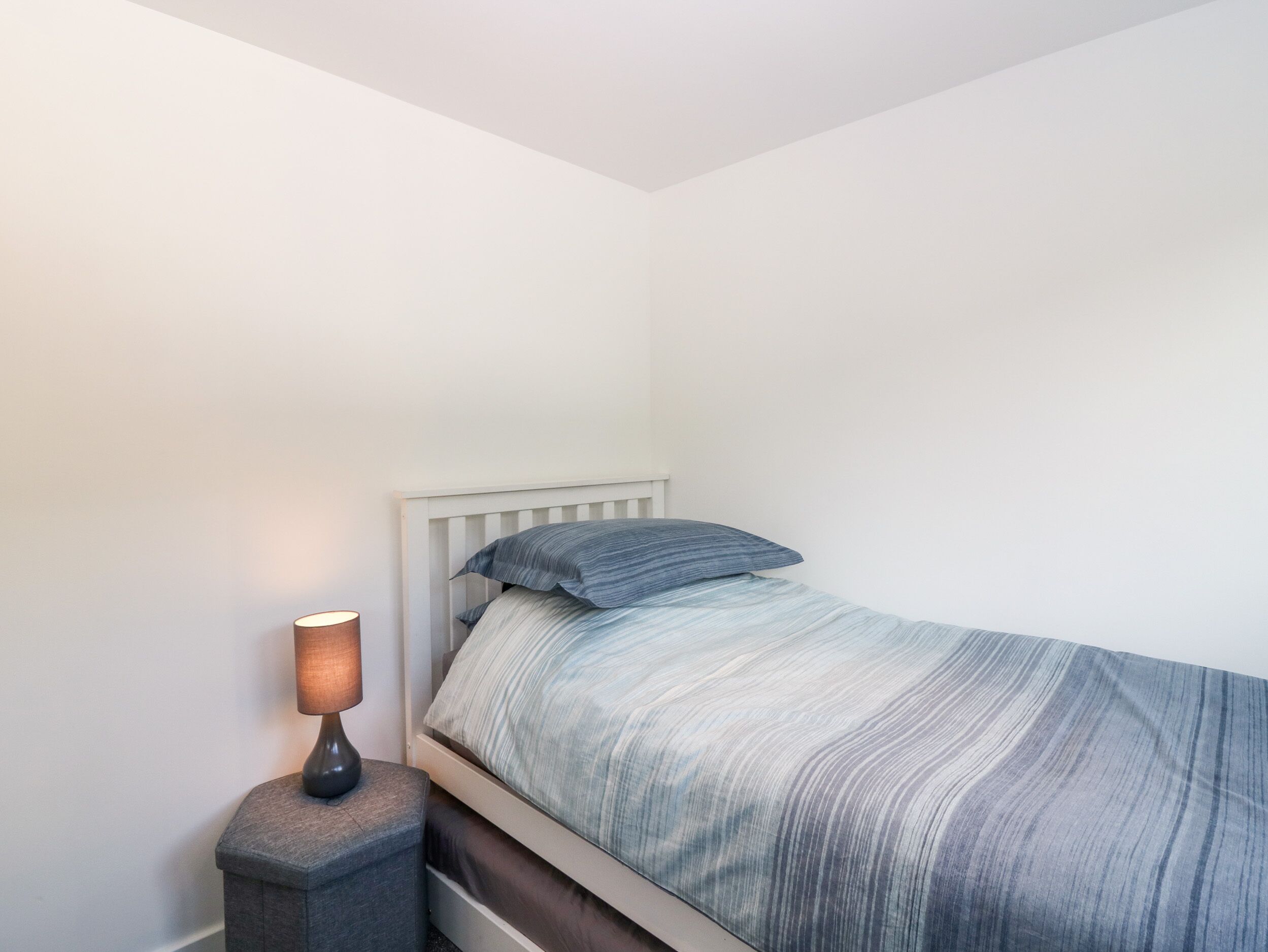 A small, neatly made bed with a striped bedspread and two pillows, a bedside lamp, and a nightstand.