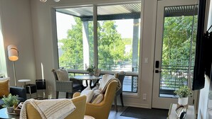 A modern living room with a sofa, armchairs, a coffee table, and a TV. There is a large window with a view of trees and a balcony. - Modern, Chic Adult ONLY Retreat On Hill w/Sunset Views Steps from Downtown  (Saugatuck)
