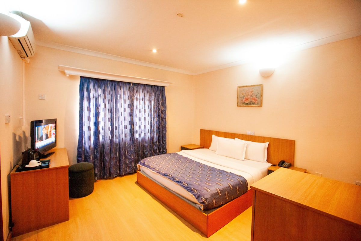 Habitación de hotel con cama, escritorio con televisor, una silla, ventana con cortinas y un cuadro en la pared.