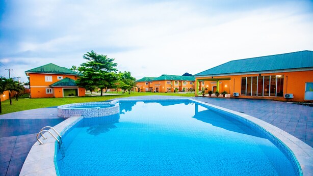 A swimming pool with a tiled edge, surrounded by a paved area and a building with a green roof. - GENESIS COTTAGE (Port Harcourt)