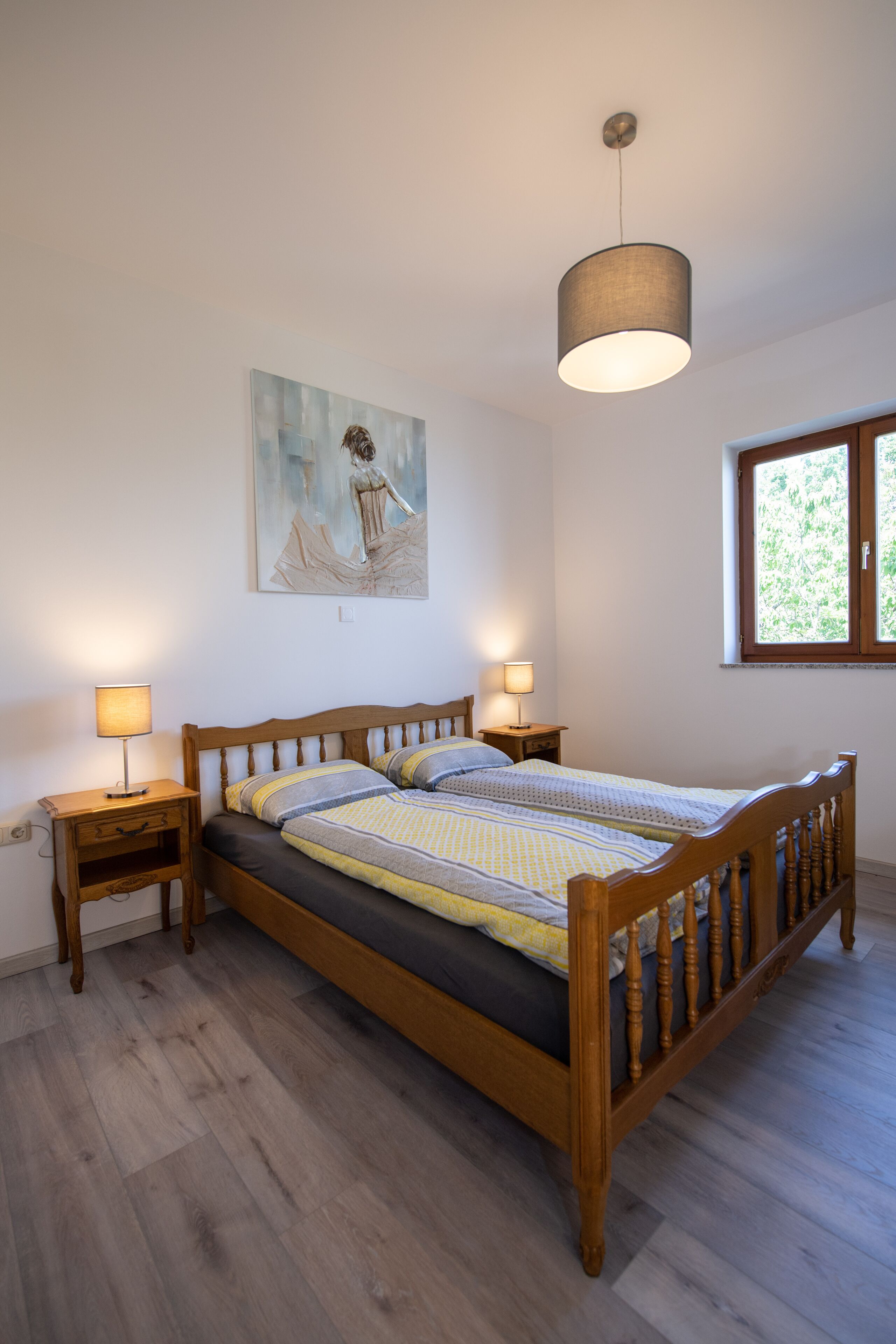 Une chambre à coucher avec un lit en bois, deux tables de chevet, une lampe suspendue, une fenêtre et un tableau au mur.