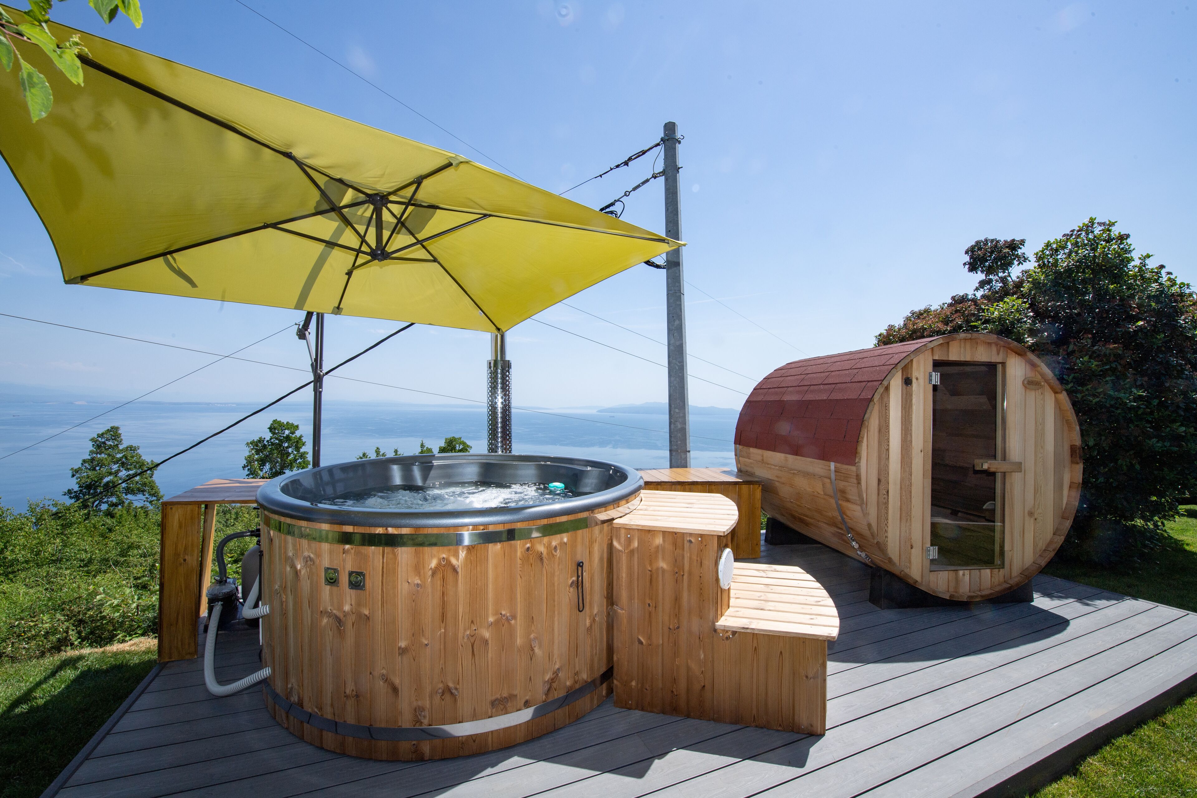 A wooden hot tub and sauna on a deck with a yellow umbrella, overlooking a scenic view.