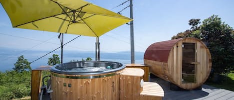 A wooden hot tub and sauna on a deck with a yellow umbrella, overlooking a scenic view.