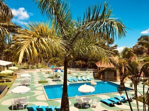 Zona de piscina con sillones de descanso, sombrillas y palmeras.