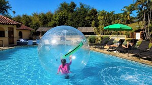 Une piscine à l’eau claire, une personne à l’intérieur d’une sphère transparente, des chaises longues et des palmiers.