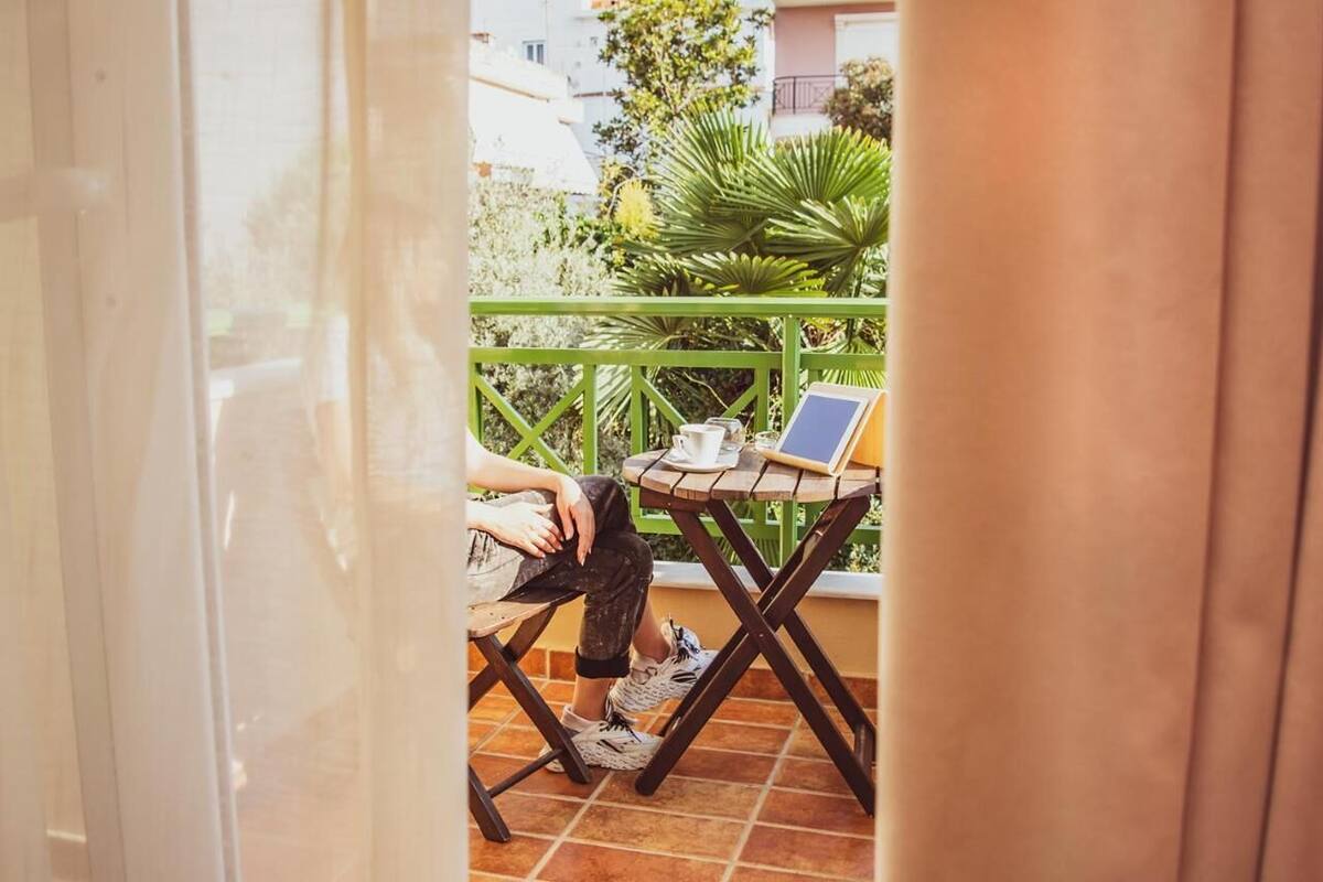 A person sitting on a chair at a small table with a laptop, a cup, and a book on a balcony with a green railing.